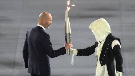 Eine mysteriöse Gestalt übergibt die Fackel an Zinedine Zidane.